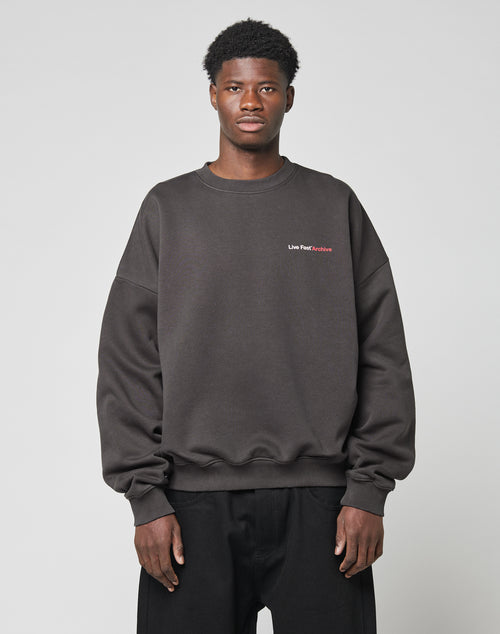 A man stands facing forward in an oversized black LFDY F*ck Sweater by LIVE FAST DIE YOUNG, made from a soft cotton-polyester blend with small white and red Live Fast Archive text on the upper left chest, paired with black pants.