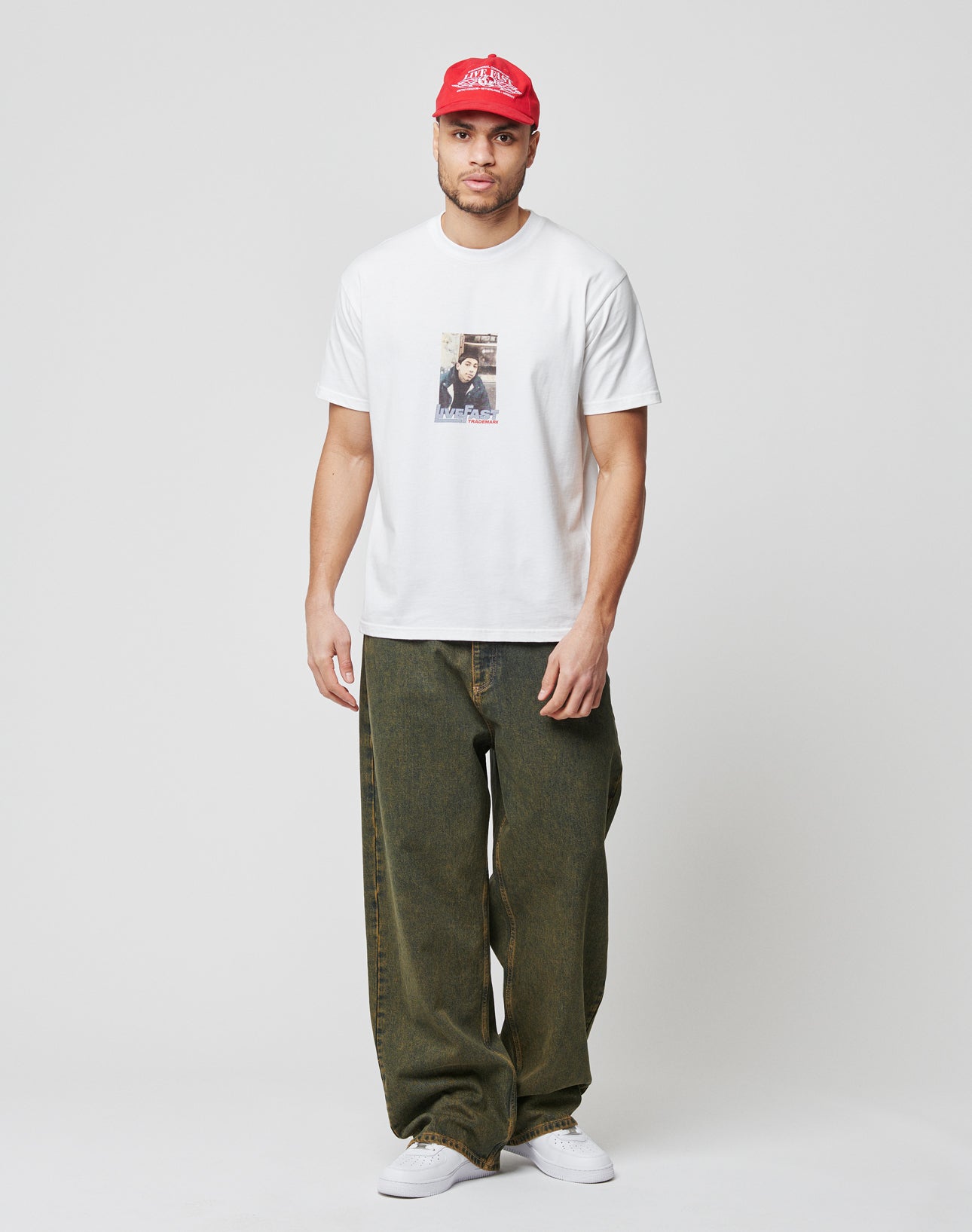 A man in a white graphic tee aus 100% Baumwolle, LFDY Baggy Denim von LIVE FAST DIE YOUNG im dunklen Grünton, weiĆen Sneakern und roter Baseballcap steht vor weiĆem Hintergrund und blickt nach vorn.