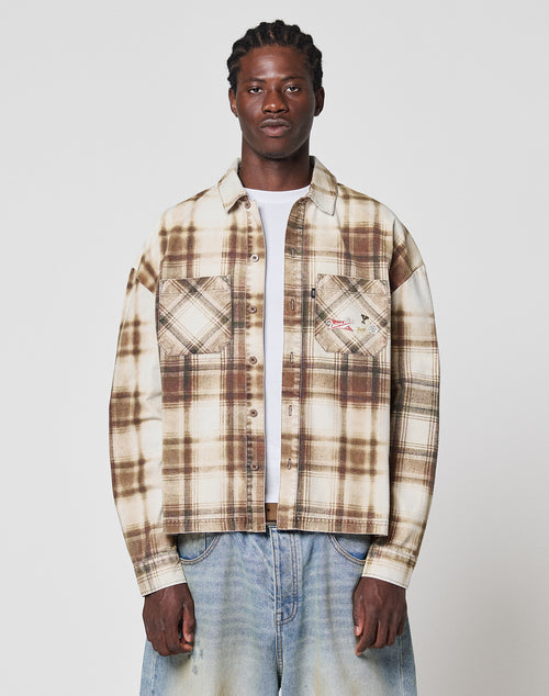 A person faces forward against a plain white background, wearing LIVE FAST DIE YOUNG’s Faded Flannel in beige and brown plaid with two chest pockets and oversize fit, paired with light blue baggy jeans—all made from soft Baumwolle.