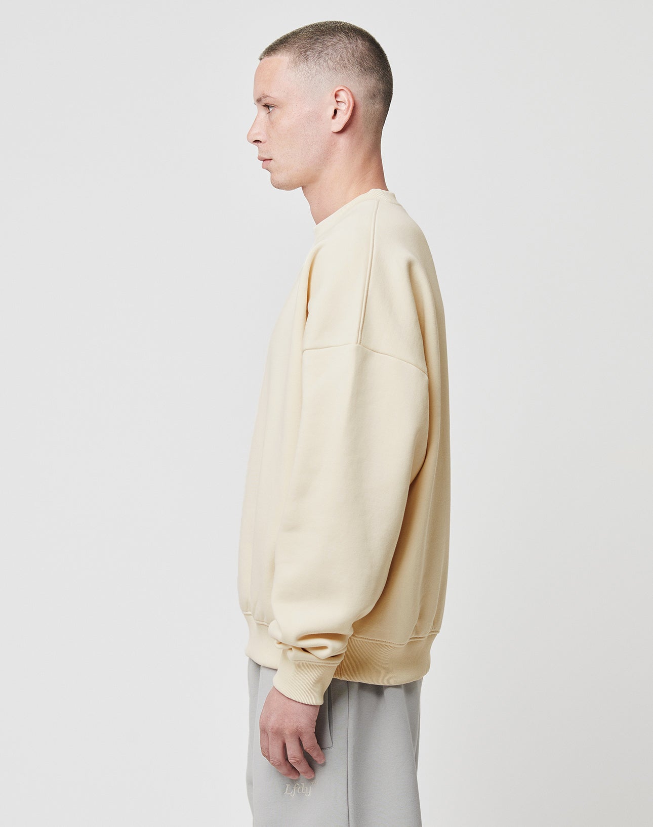 A person with short hair faces left, wearing the LIVE FAST DIE YOUNG Basic Blank Sweater in beige and light gray pants, standing against a plain white background.
