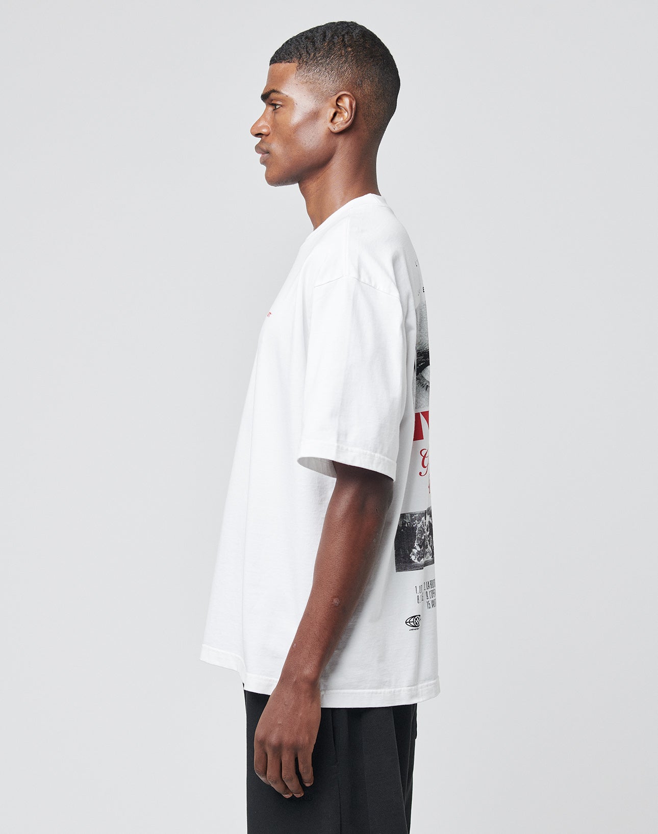 A young man stands in profile against a light gray background, wearing black pants and the LIVE FAST DIE YOUNG LFDY Cinema Teeāan Oversize Fit white T-shirt made of 100% Baumwolle with front and back prints featuring text and images.