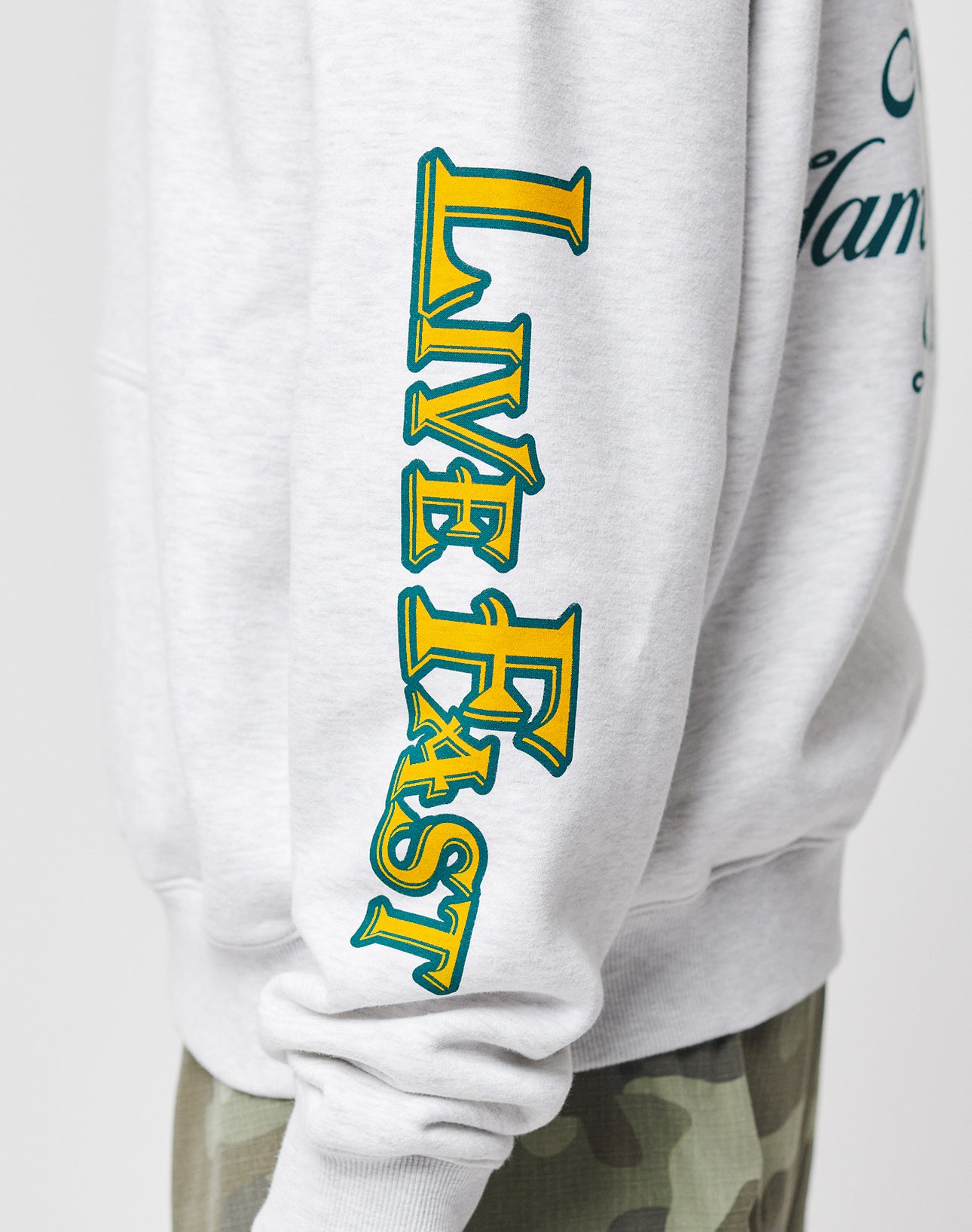A person wearing a light gray Rhine Area Hooded sweatshirt by LIVE FAST DIE YOUNG with bold yellow and green LIVE FAST lettering on the sleeve, paired with camouflage pants.