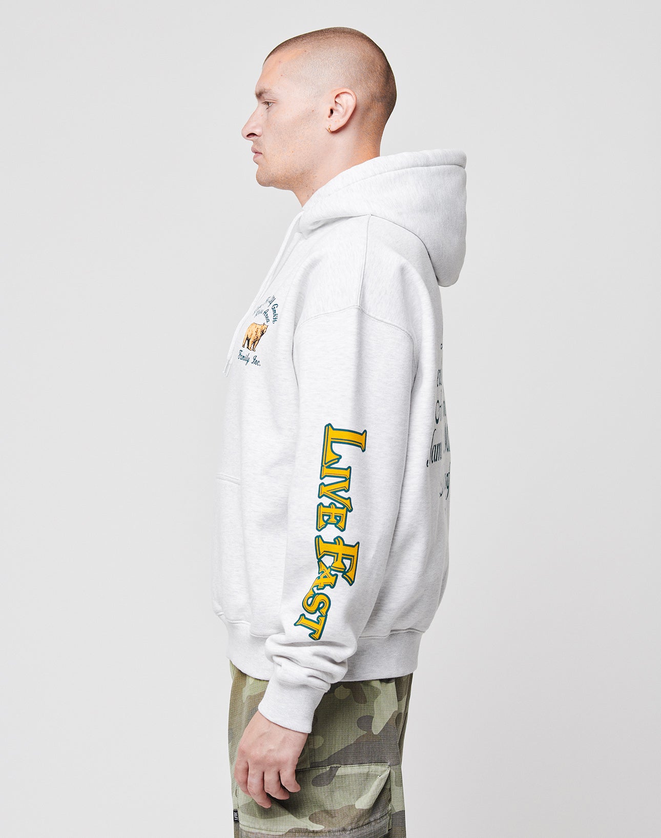 A person with short hair wears an oversize light gray Rhine Area Hooded sweatshirt by LIVE FAST DIE YOUNG, featuring yellow and green sleeve text, paired with green camo pants, standing in profile against a plain background.