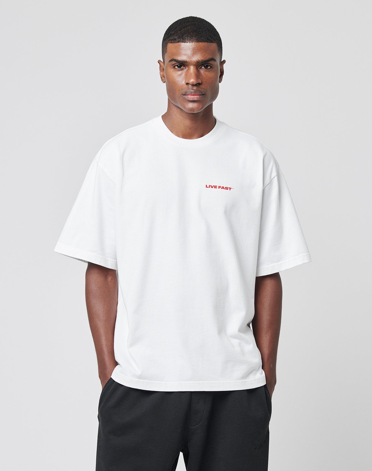 A man stands against a plain light gray background with his hands in his pockets, wearing the LIVE FAST DIE YOUNG LFDY Cinema Teeāan oversize fit 100% cotton white t-shirt with LIVE FAST. printed in small red letters on the left chest.