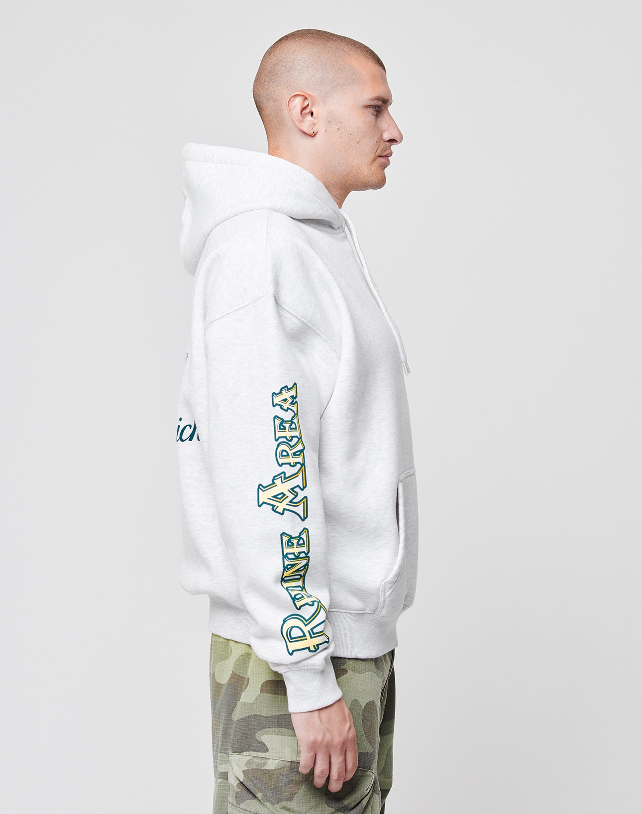 A person in profile wears an oversize light gray LIVE FAST DIE YOUNG Rhine Area Hooded with BAY AREA in green text on the sleeve and camouflage cargo shorts, standing against a plain white background.