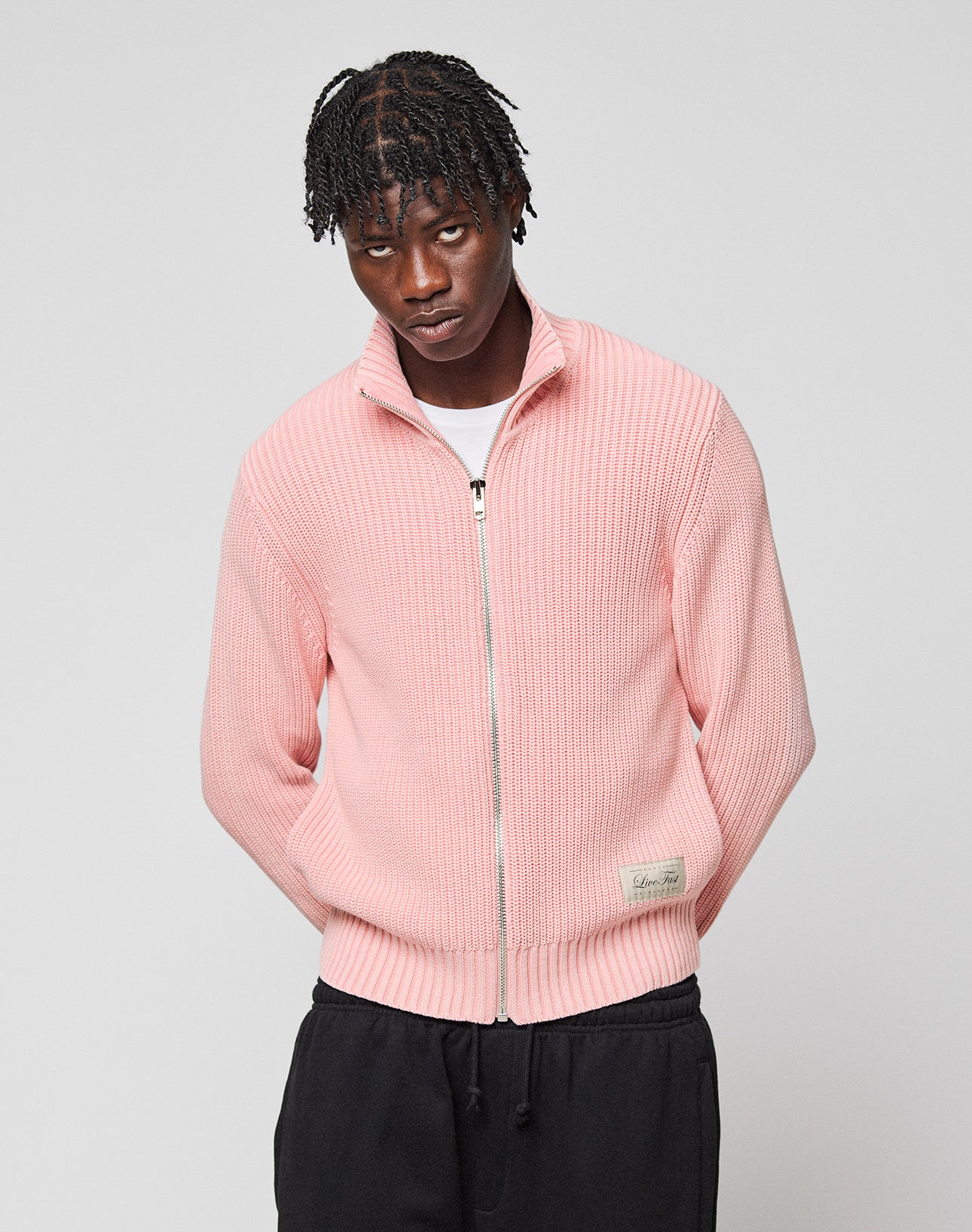 A man with braided hair wears the LIVE FAST DIE YOUNG LFDY Knit Jacket in light pink over a white t-shirt and black pants, standing against a plain light gray background.