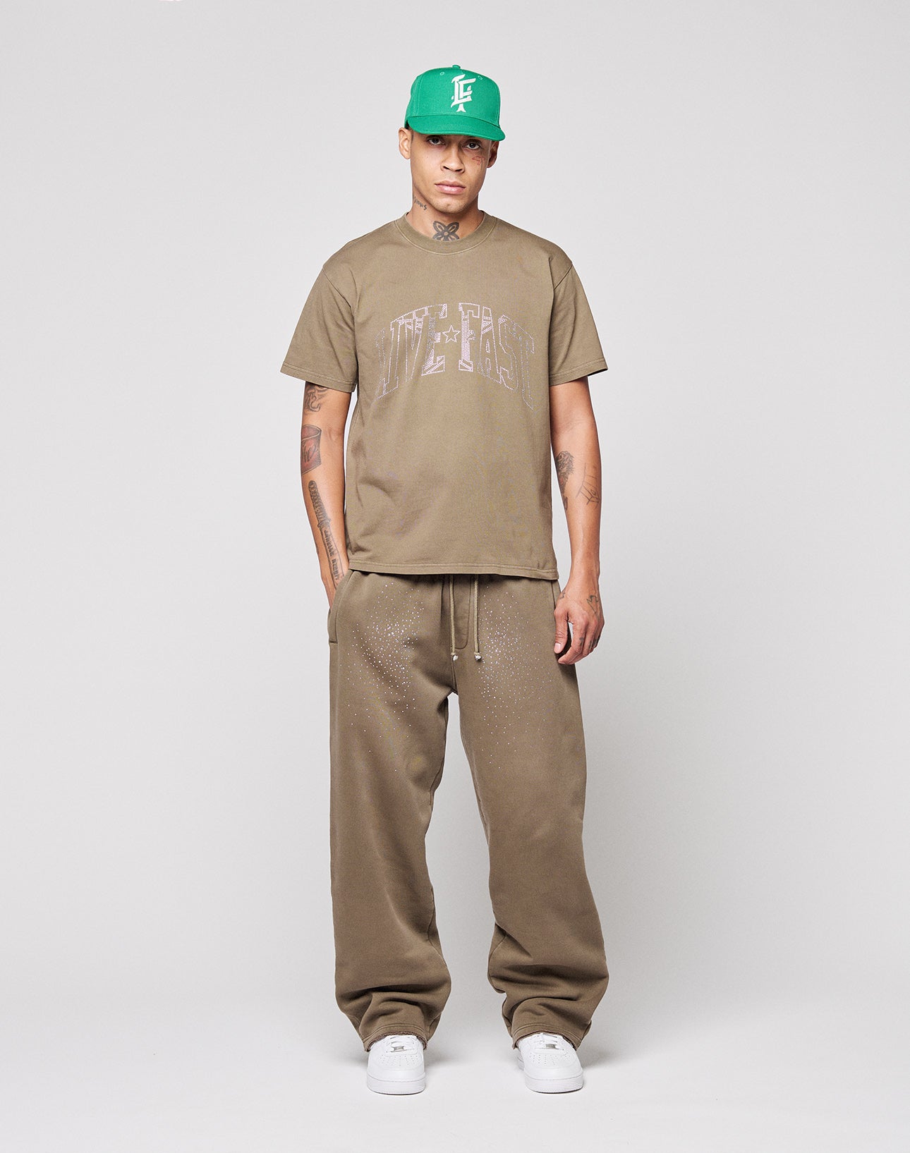 A person stands facing forward, wearing a green baseball cap, LIVE FAST DIE YOUNG's Live Fast Rhinestone Tee in brown, matching brown sweatpants, and white sneakers against a light gray background.