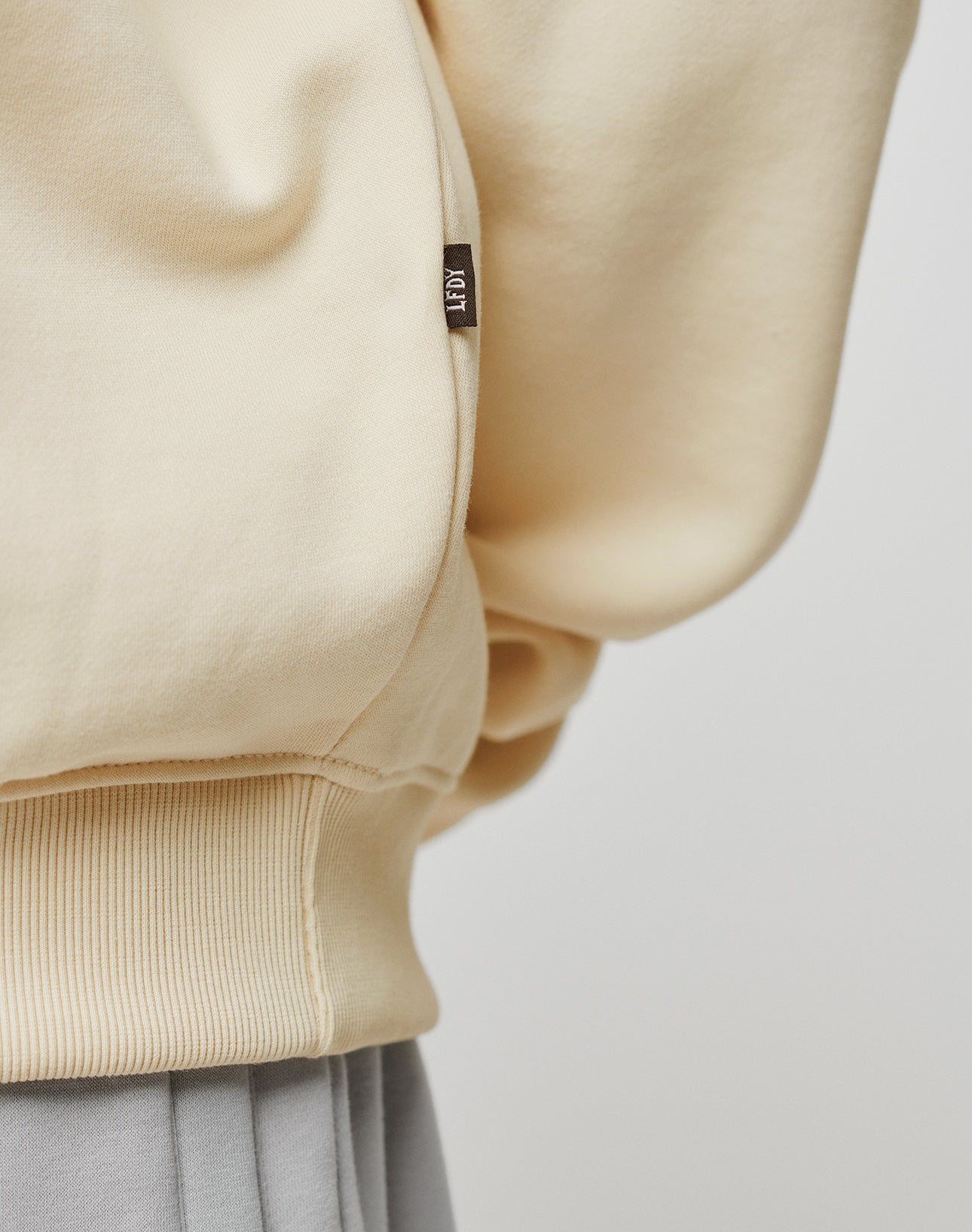 Close-up of the cream-colored Basic Blank Sweater by LIVE FAST DIE YOUNG, showing ribbed hem, detailed topstitching, and a small brown tag with white text. Worn over light grey pants against a plain light background.