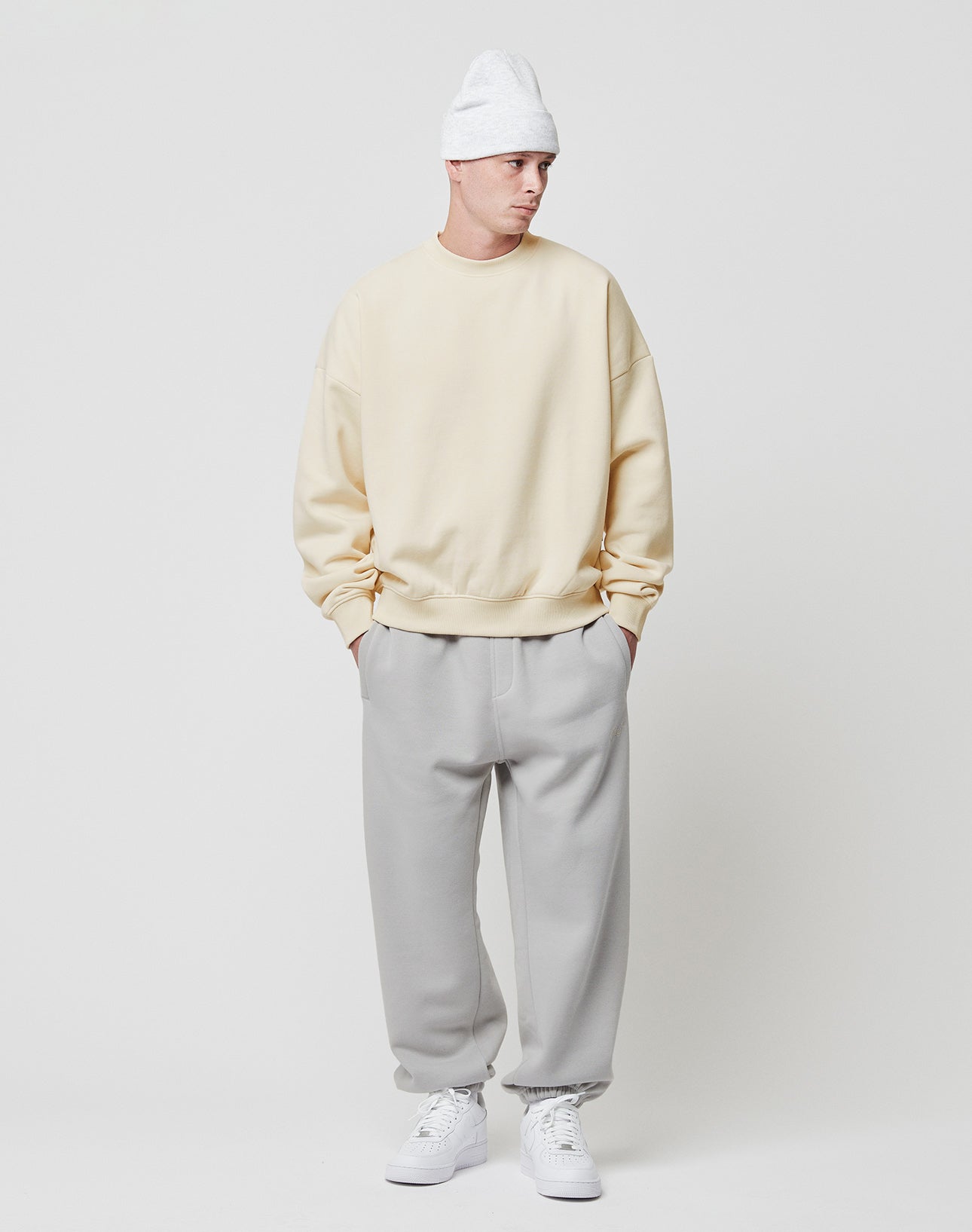 A person stands against a white background, wearing a LIVE FAST DIE YOUNG Basic Blank Sweater in cream, light gray sweatpants, white sneakers, and a white knit beanie—hands in pockets and gazing slightly to the side.