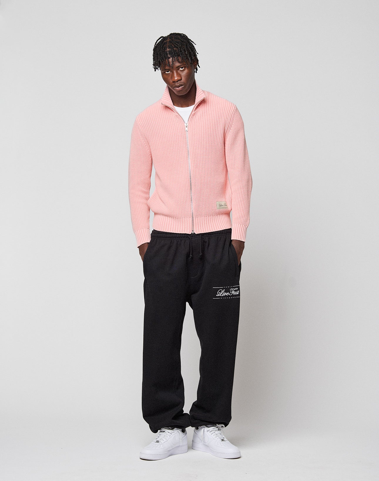 A man with short, twisted hair and a neutral expression stands against a light background wearing the LIVE FAST DIE YOUNG LFDY Knit Jacket in light pink with Stehkragen, black sweatpants with white text on the left, and white sneakers.