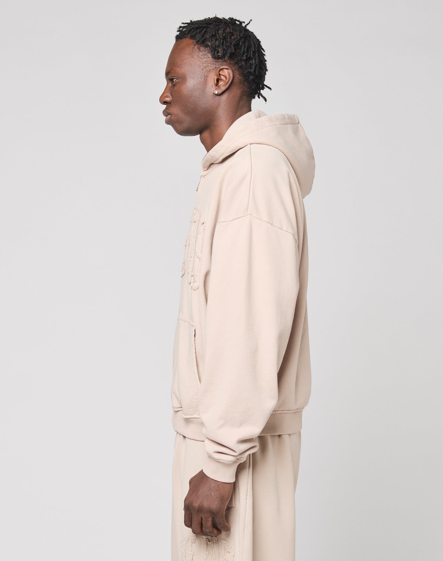 A person with short, braided hair stands in profile against a plain light background, wearing the LIVE FAST DIE YOUNG Ripped College Hooded Zip—an oversized beige sweatshirt made from soft Baumwolle.