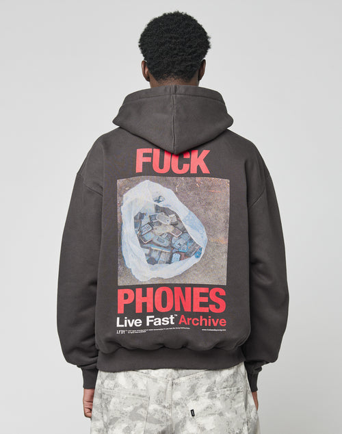 A person wears the LIVE FAST DIE YOUNG LFDY F*ck Phones Hooded, an oversize dark hoodie made of soft cotton and polyester, featuring bold red and white āF*CK PHONES Live Fast Archiveā text and a graphic of old phones in a plastic bag on the back.