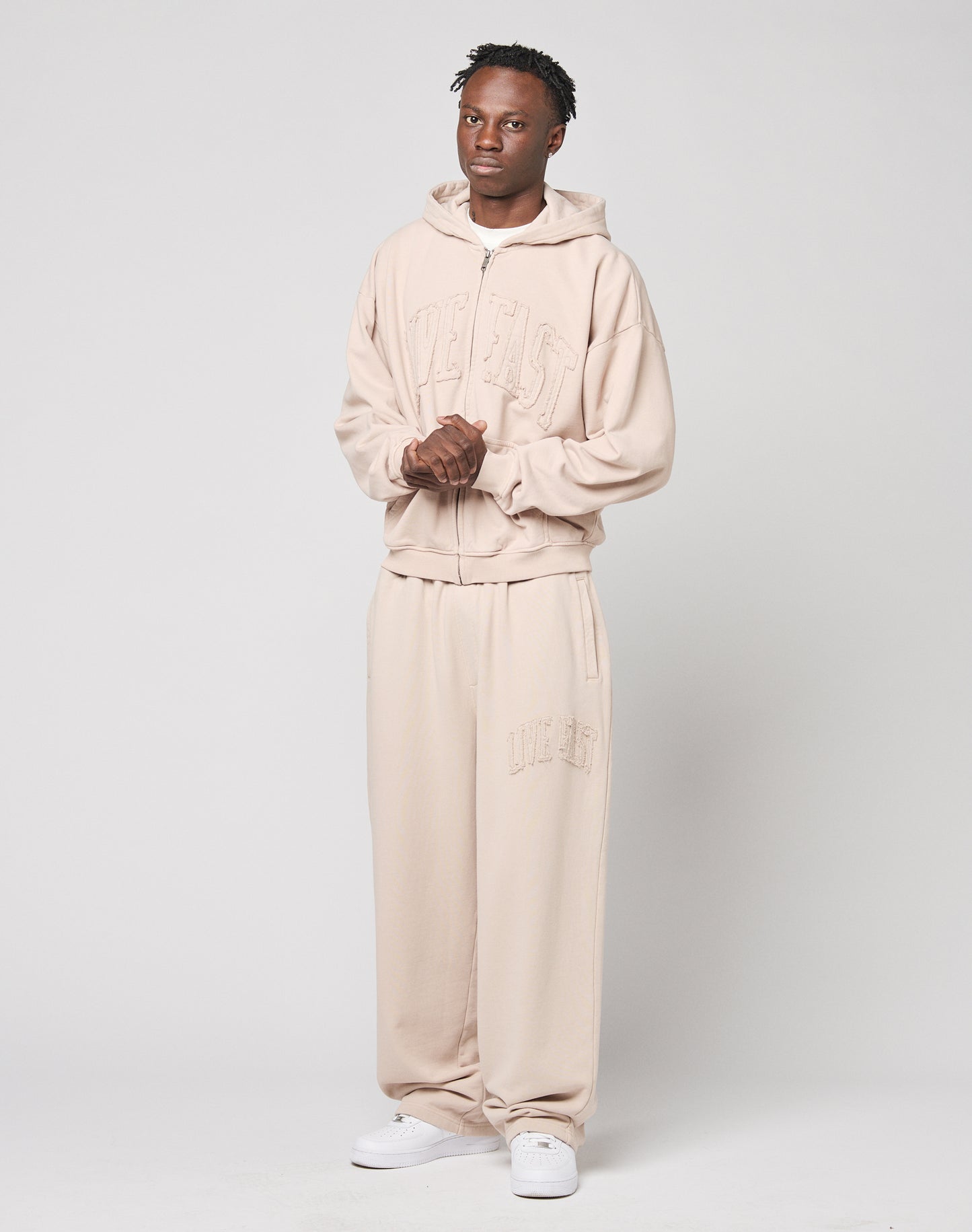 A person stands against a plain background wearing LIVE FAST DIE YOUNG’s Ripped College Wide Jogger in light beige cotton, paired with a matching wide hoodie and white sneakers. Embossed text adds detail as their hands are clasped in front.