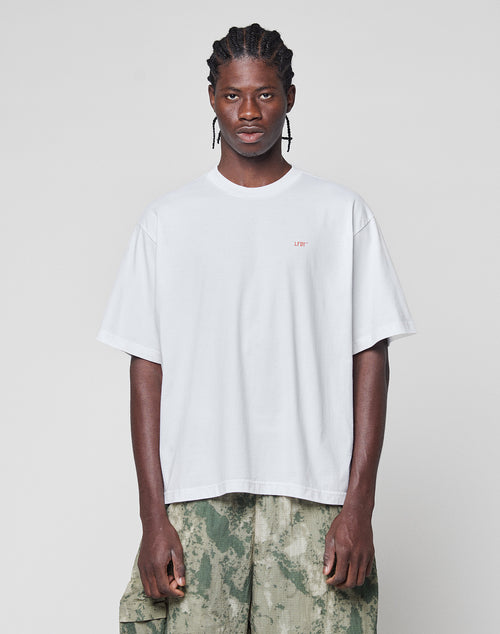 A person with braided hair wears the LIVE FAST DIE YOUNG Live Fast Vegas Tee, a loose white T-shirt with a small red logo on the left chest, and green camo cargo shorts, standing against a plain light gray background.