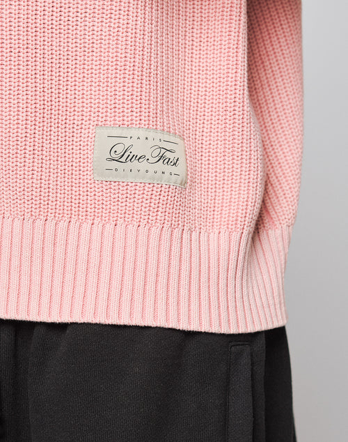 A close-up of the LFDY Knit Sweater by LIVE FAST DIE YOUNG in light pink, with a fabric label and trendy dropped shoulders, worn above black pants and featuring an oversized fit.
