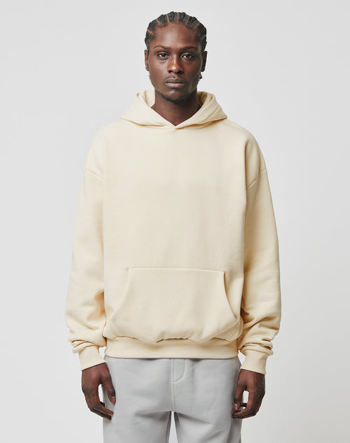 A person with braided hair wears the LIVE FAST DIE YOUNG Basic Blank Hooded cream-colored, oversize hoodie made from soft cotton and polyester, paired with light gray pants, standing against a plain white background.