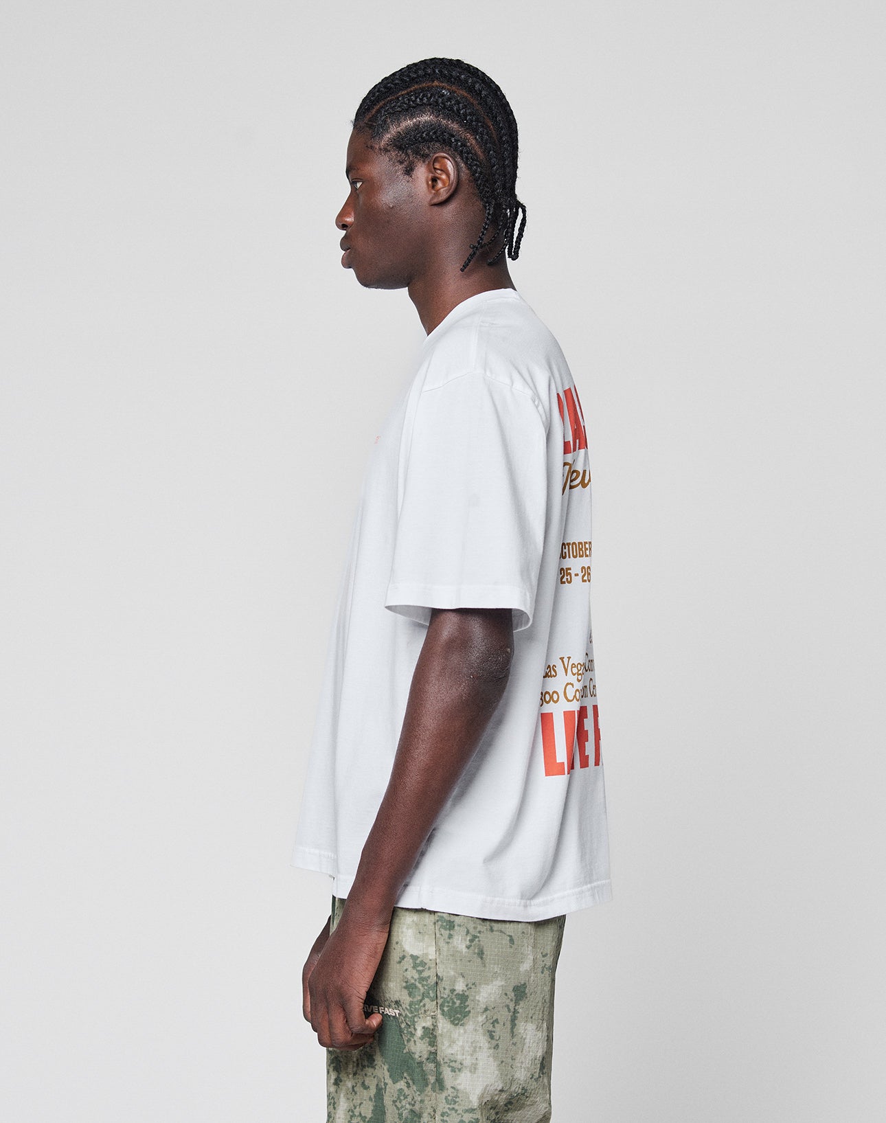 A person with braided hair stands in profile wearing the LIVE FAST DIE YOUNG Live Fast Vegas Tee, an oversized white graphic T-shirt with front and back prints, paired with green camouflage pants against a plain light gray background.
