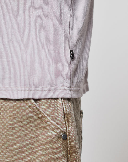 Close-up of a person wearing the LFDY Rib Longsleeve by LIVE FAST DIE YOUNG in light mauve with an oversize fit and black hem tag, paired with light brown denim pants, highlighting the ribbed texture, topstitching, and clothing details.