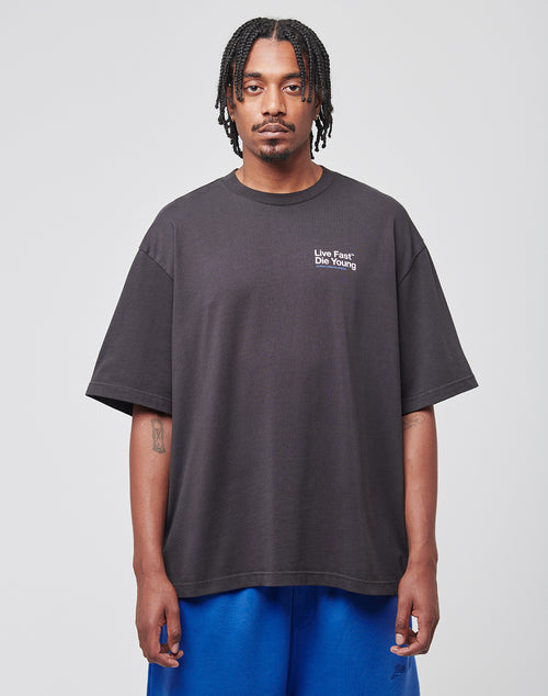 A man with braids faces forward, wearing the LIVE FAST DIE YOUNG Live Fast NY Tee in black and blue shorts, against a plain light gray background.