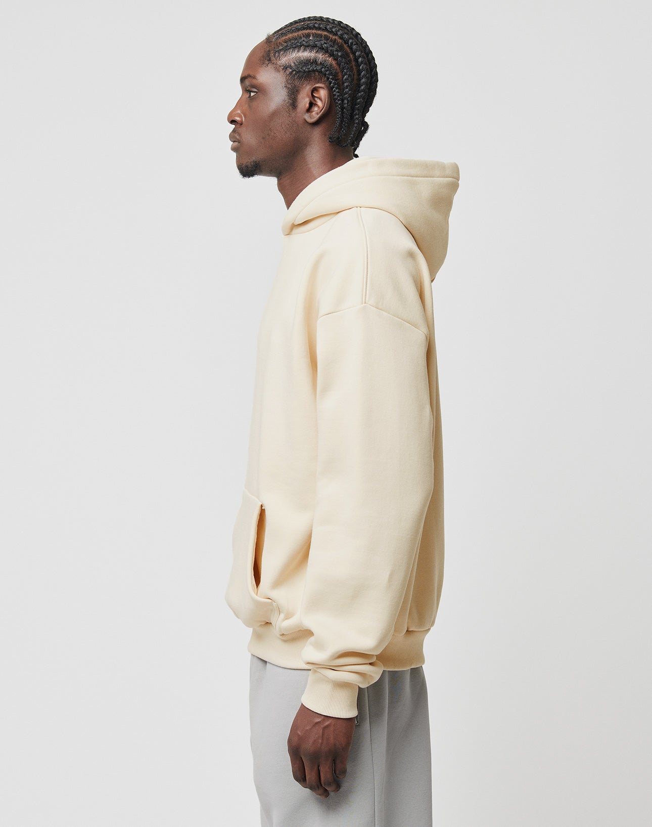 A man with braided hair stands in profile, wearing the LIVE FAST DIE YOUNG Basic Blank Hooded in light beige (oversize fit, Baumwolle and Polyester), paired with light gray pants against a plain white background.