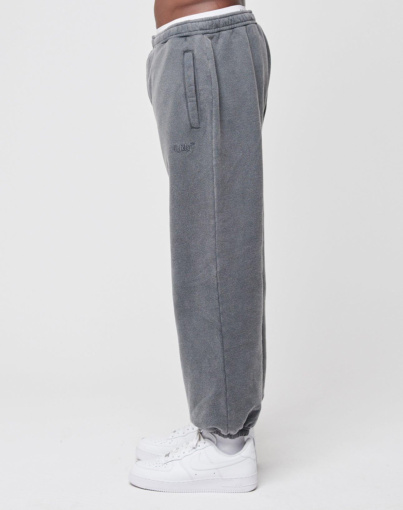 A person, shown from the waist down, wears LIVE FAST DIE YOUNG’s gray Loose Jogger sweatpants with an Elastik-Bund and white sneakers, standing sideways against a plain white background.
