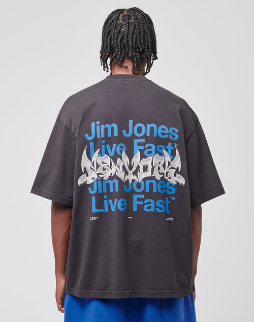 A person with braided hair, facing away from the camera, wears the LIVE FAST DIE YOUNG Live Fast NY Tee—an oversized cotton T-shirt with a blue Jim Jones front print and bold graffiti-style New York back print.
