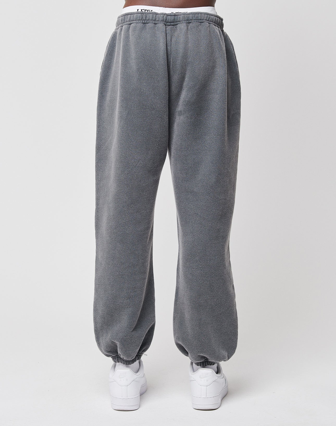 A person wears LIVE FAST DIE YOUNG Loose Jogger sweatpants with elastic waistband in gray cotton and white sneakers, standing against a plain white background.