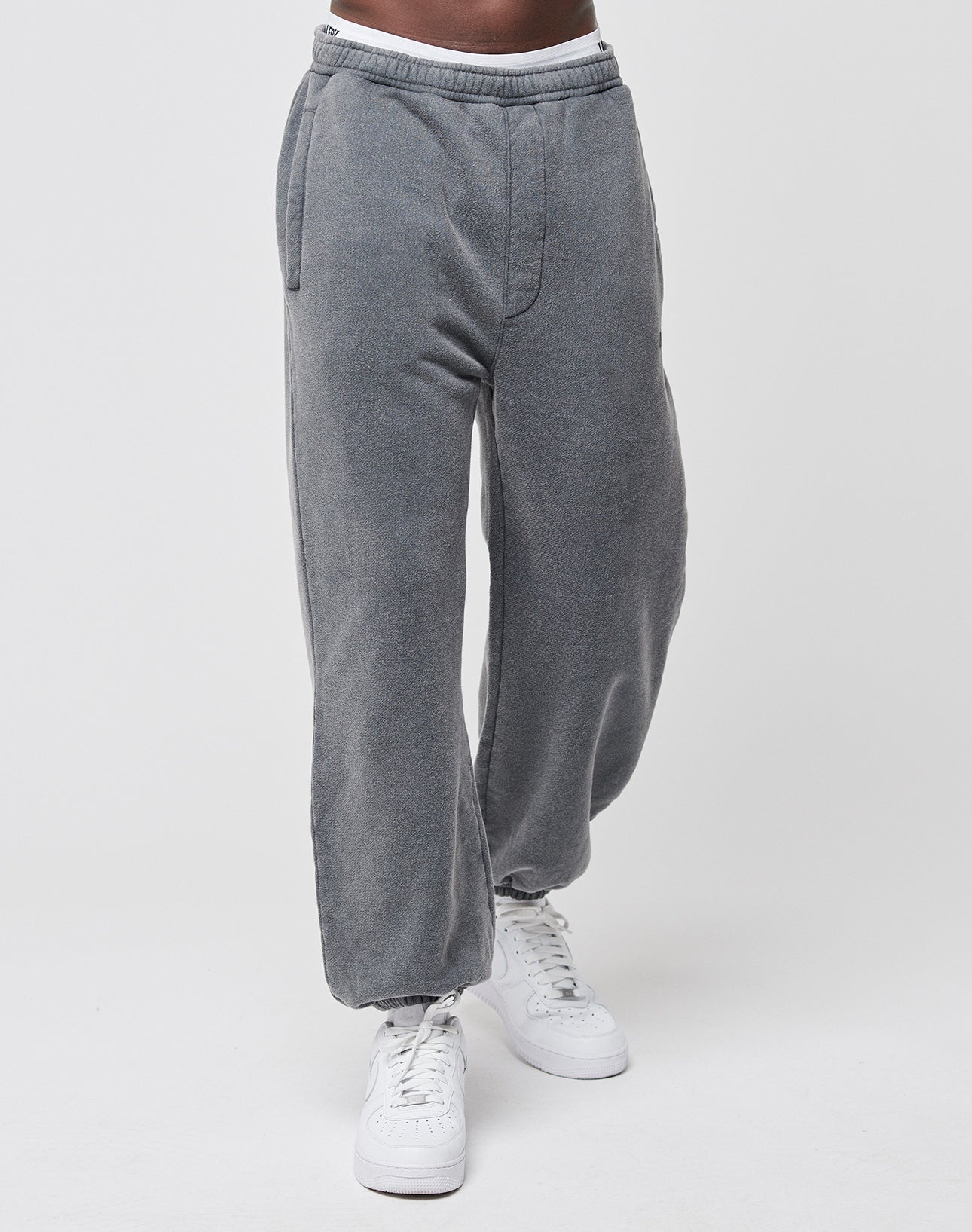 A person stands against a white background wearing LIVE FAST DIE YOUNG Loose Jogger sweatpants—gray, cotton, loose fit with elastic waistband and side pockets—paired with white sneakers. Only the lower body is visible.