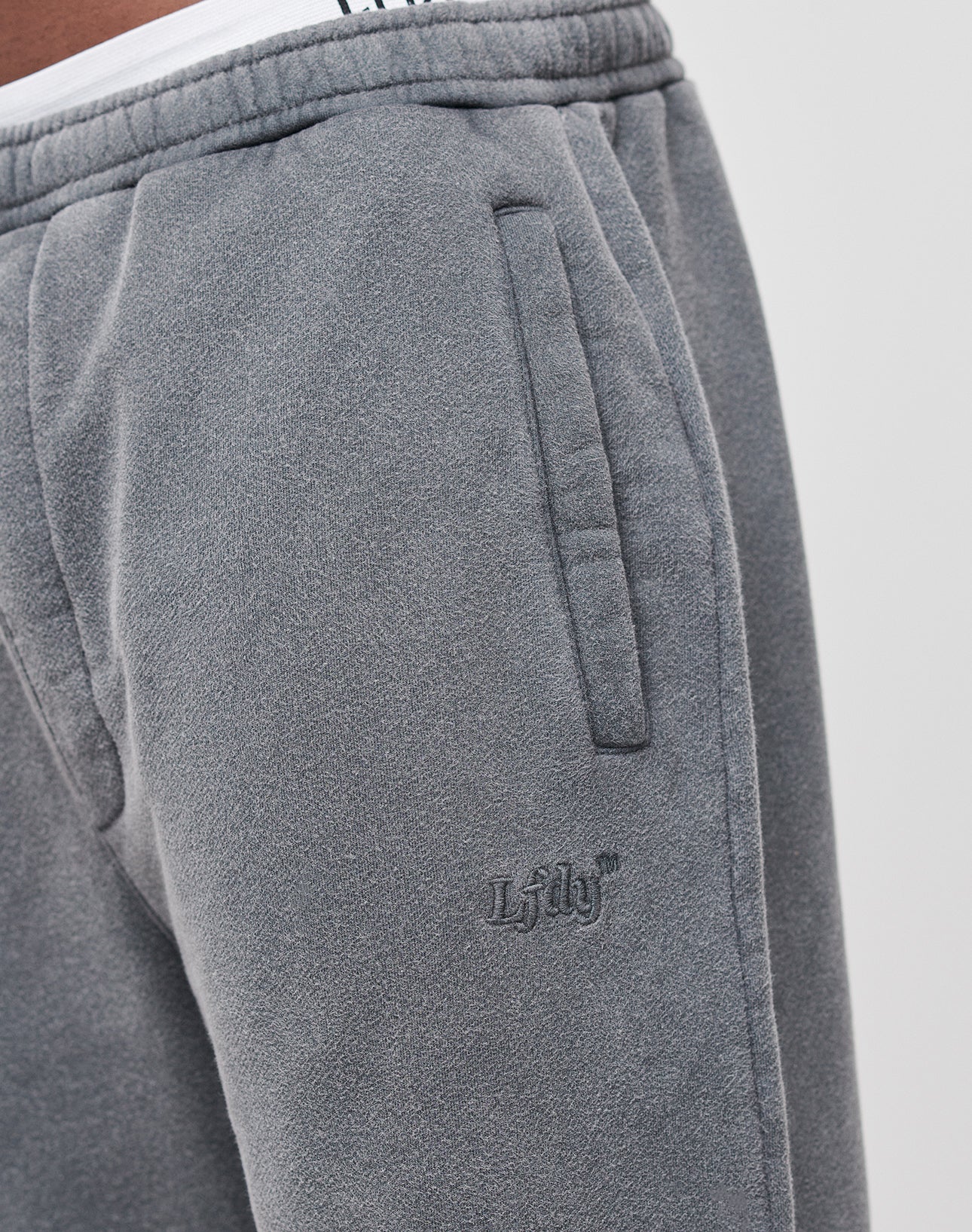 Close-up of a person wearing LIVE FAST DIE YOUNG Loose Jogger in gray fleece with side pockets and Lily embroidery. The visible Elastik-Bund waistband and Loose Fit offer ultimate comfort and relaxed style.