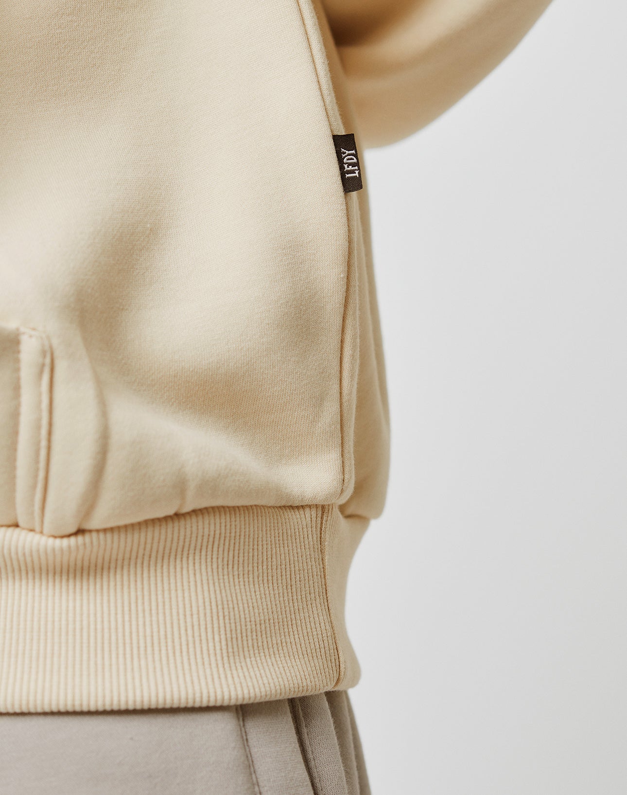Close-up of the lower side of a beige Basic Blank Hooded by LIVE FAST DIE YOUNG in an oversize fit, featuring a ribbed hem, side seam, and small black tag with white LESY text. Made from soft Baumwolle. Background is plain and light-colored.