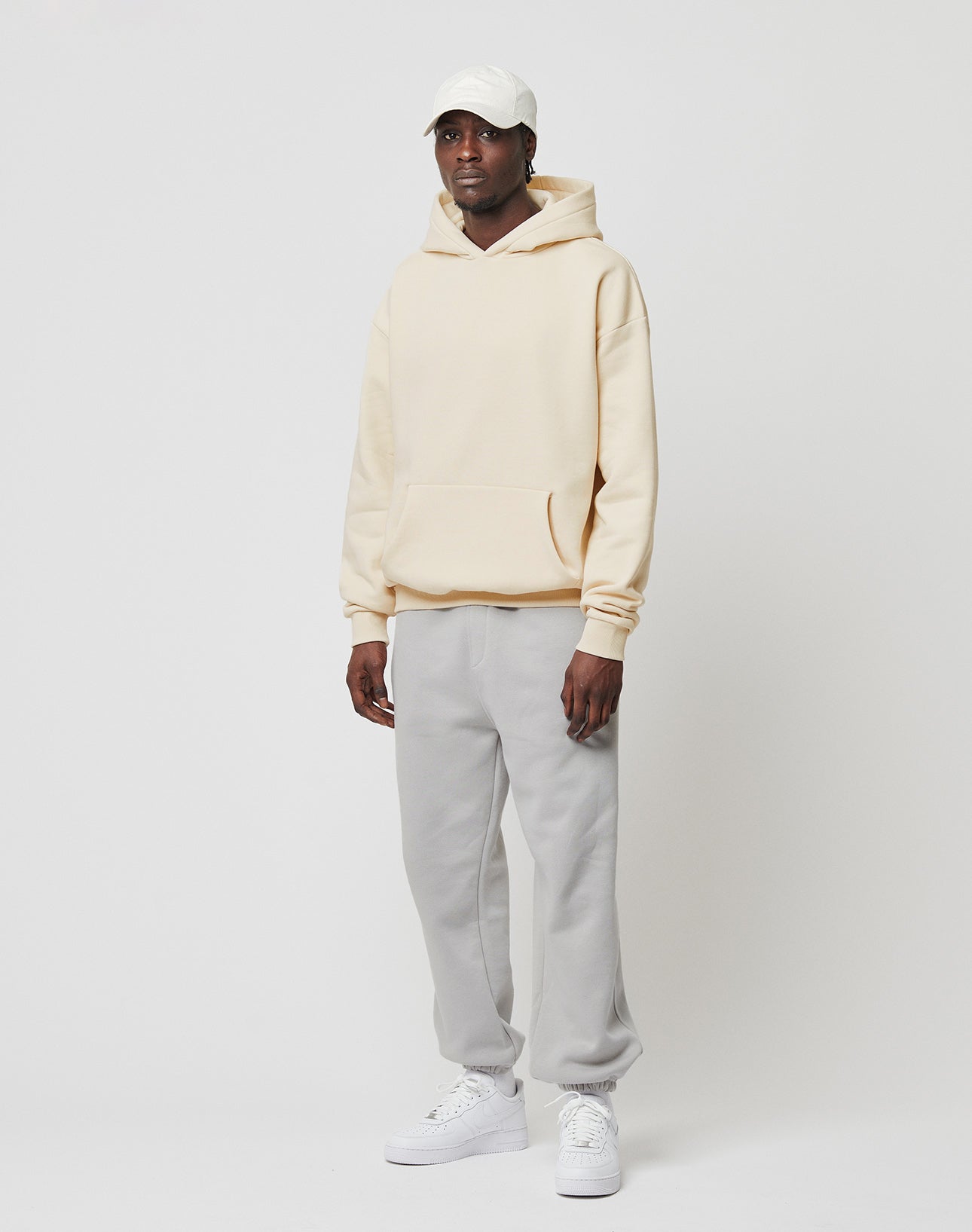A person wears an oversized cream Basic Blank Hooded by LIVE FAST DIE YOUNG, light gray sweatpants, white sneakers, and a white cap, standing against a plain light background with a relaxed posture, gazing slightly to the side.
