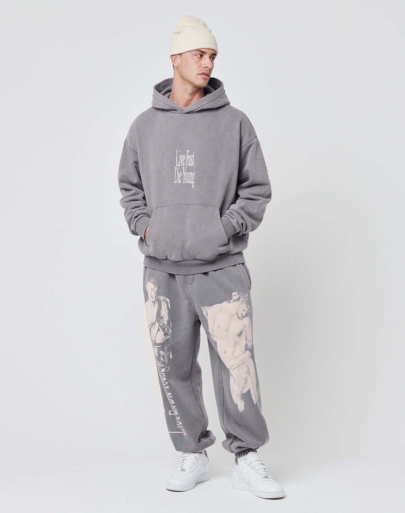 A person in an oversized gray LFDY Stitched Hooded sweatshirt by LIVE FAST DIE YOUNG, styled with matching gray sweatpants featuring classical art prints and white sneakers, stands against a plain backdrop. The look is crafted from soft cotton and polyester.