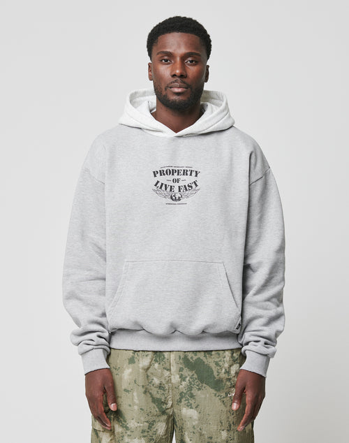 A man in an oversized light gray Live Fast Property Hooded by LIVE FAST DIE YOUNG with chest frontprint and Kapuze, paired with camo pants, stands facing the camera against a plain light background.