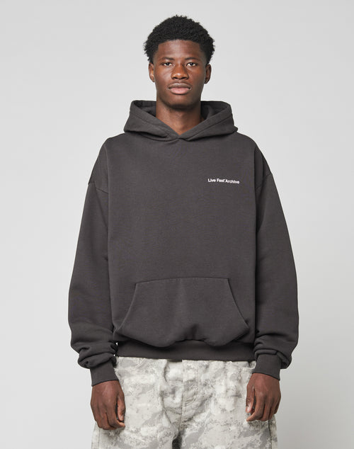 A young man with short, curly hair wears the LFDY Middlefinger Hooded by LIVE FAST DIE YOUNGāa dark, oversize-fit hoodie made from a soft polyester-cotton mixāand light gray camo pants, standing against a plain background with a neutral expression.