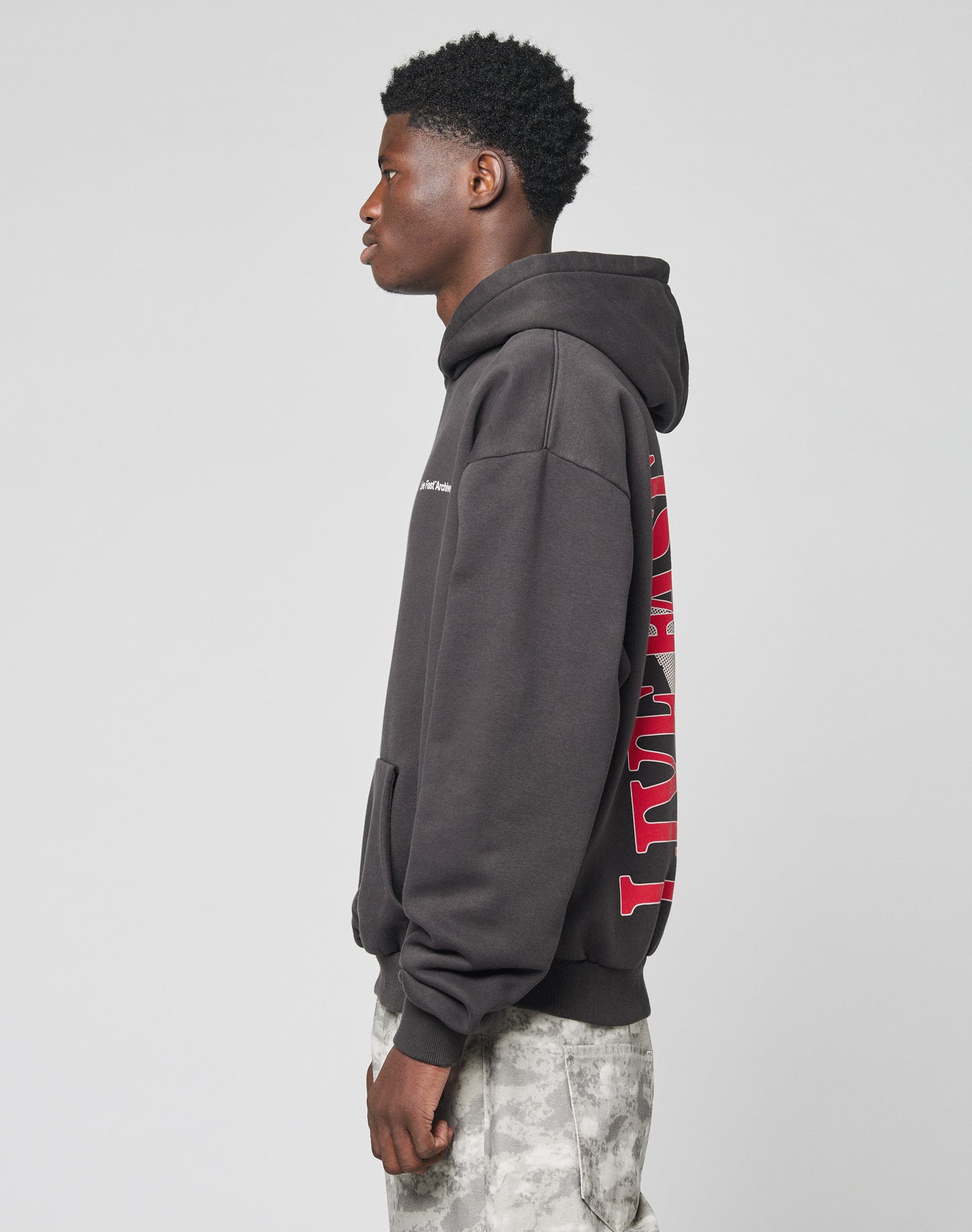 A young man stands in profile wearing the LIVE FAST DIE YOUNG LFDY Middlefinger Hooded, an oversize dark hoodie with red back text, paired with light gray patterned pants made from soft Baumwolle, against a plain light background.