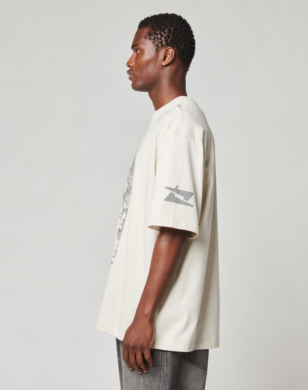 A man stands in profile against a plain light gray background, wearing the LIVE FAST DIE YOUNG LFDY Window Tee—an oversize, off-white 100% Baumwolle shirt with reflective sleeve and logo prints—paired with faded gray jeans.