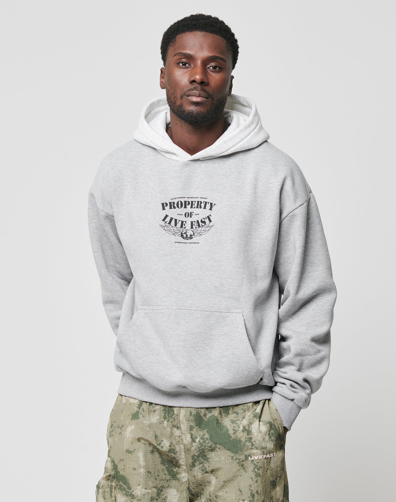 A man wearing the LIVE FAST DIE YOUNG Live Fast Property Hooded sweatshirt in light gray with front print, paired with green camo oversized pants, stands against a plain white background.