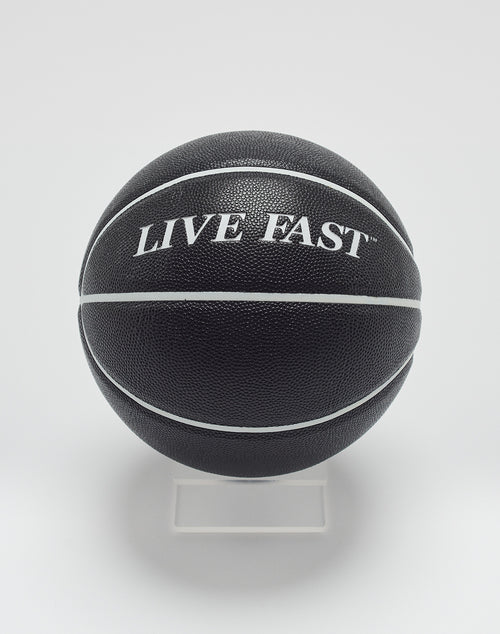 The Live Fast Basketball by LIVE FAST DIE YOUNG features a black design with white stripes, deep grooves, and LIVE FAST branding in hygroscopic PU, displayed on a clear stand against a white background.