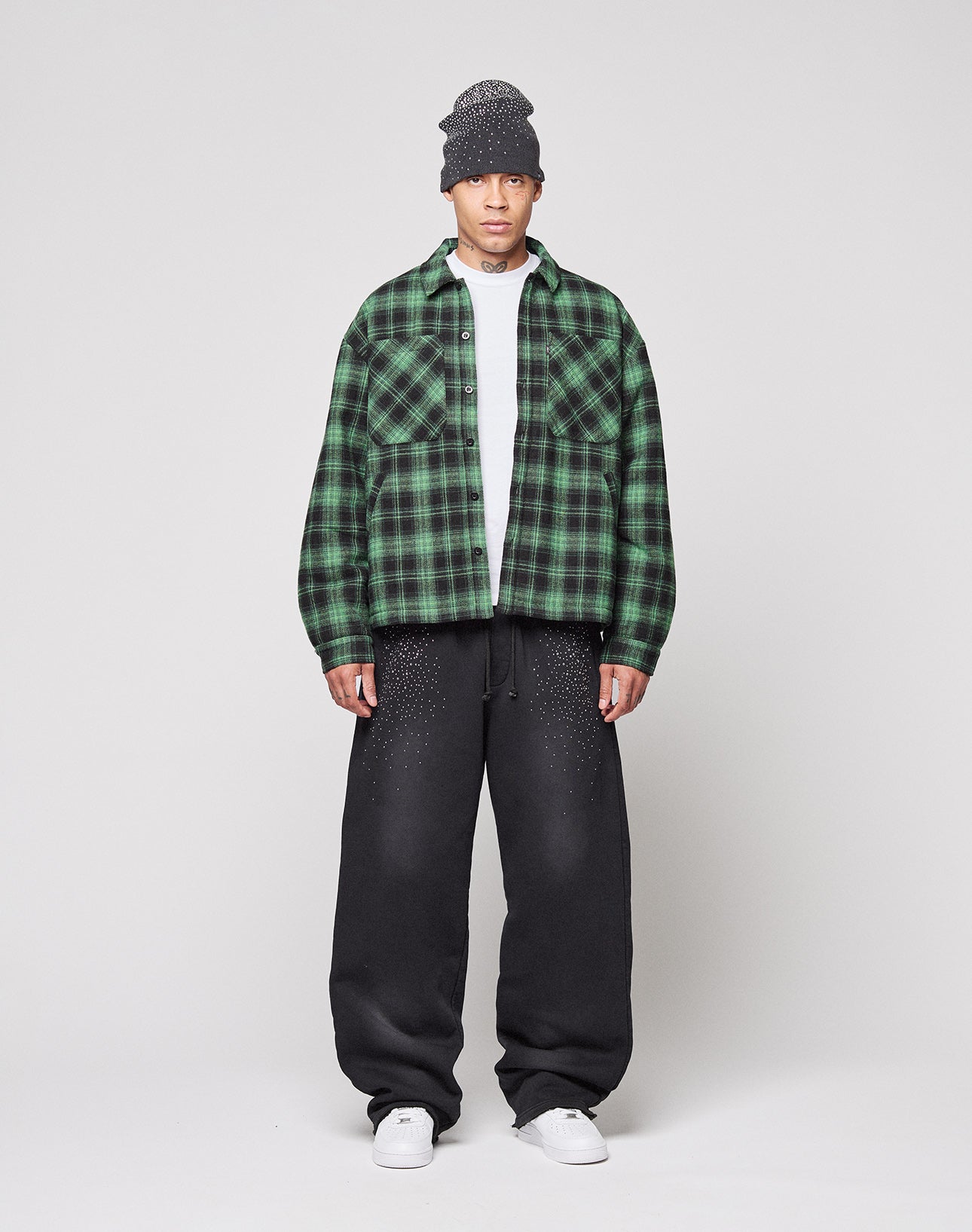 A person stands against a plain light background wearing a gray knit beanie, the LIVE FAST DIE YOUNG LFDY Padded Flannel in green and black plaid over a white shirt, black baggy pants with small white dots, and white sneakers.