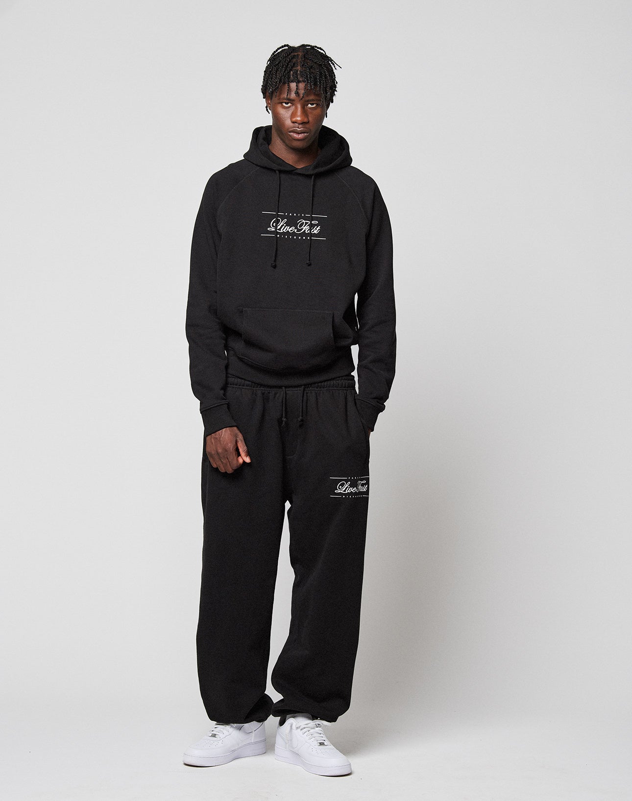 A person with short dreadlocks and a neutral expression stands against a plain light background, wearing a black LIVE FAST DIE YOUNG LFDY Embroidery Hooded hoodie and matching black sweatpants with white text, plus white sneakers.