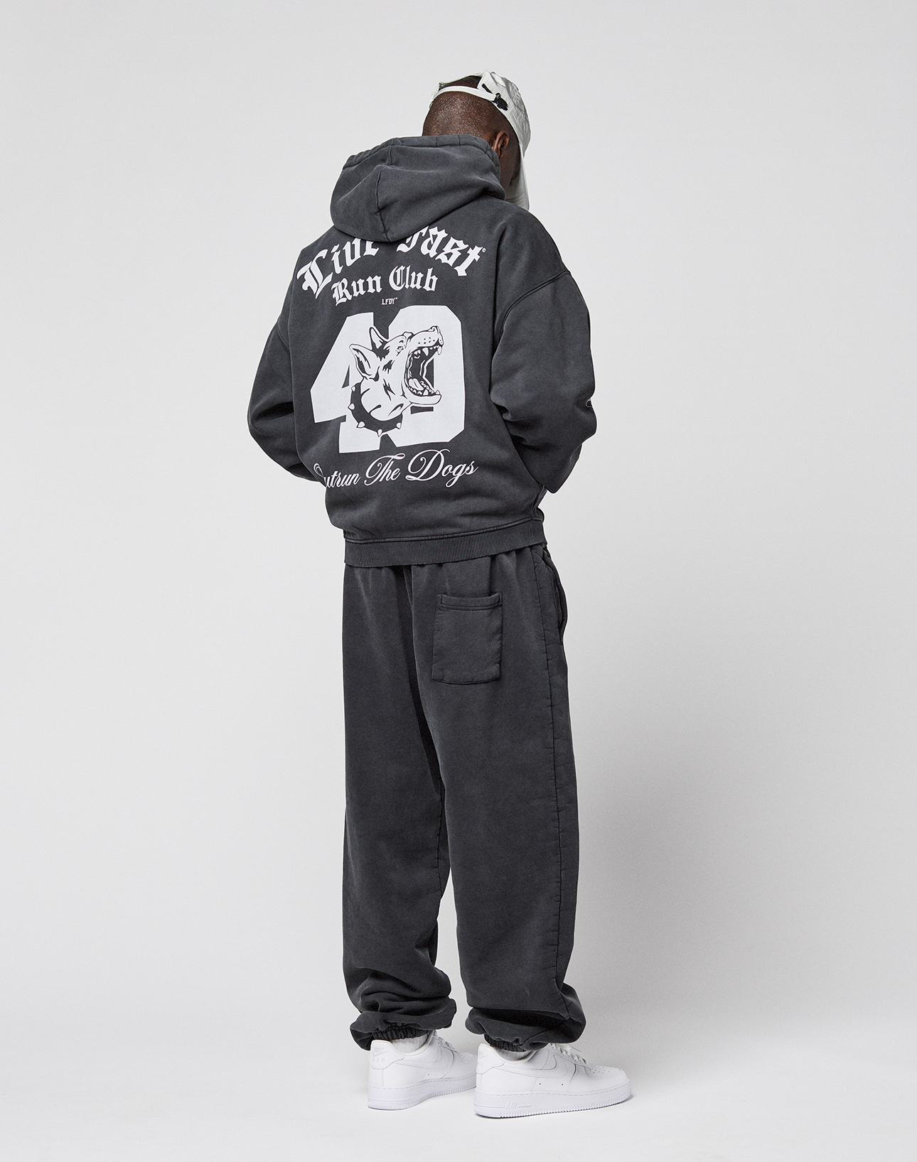 A person faces away, wearing the LIVE FAST DIE YOUNG LFDY Run Club Hooded Zip and sweatpants with a large dog head graphic and "True Issue Run Club, Down For Dogs" on the back. They pair it with white sneakers, a light cap, and an Ćrmelprint.