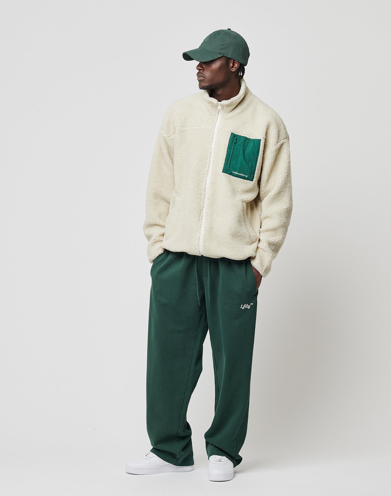 A man stands against a plain white background, looking to the side with hands in his pockets, wearing the LIVE FAST DIE YOUNG Basic Teddy Jacket, a cream fleece with nylon collar and green chest pocket, plus a green cap, green pants, and white sneakers.