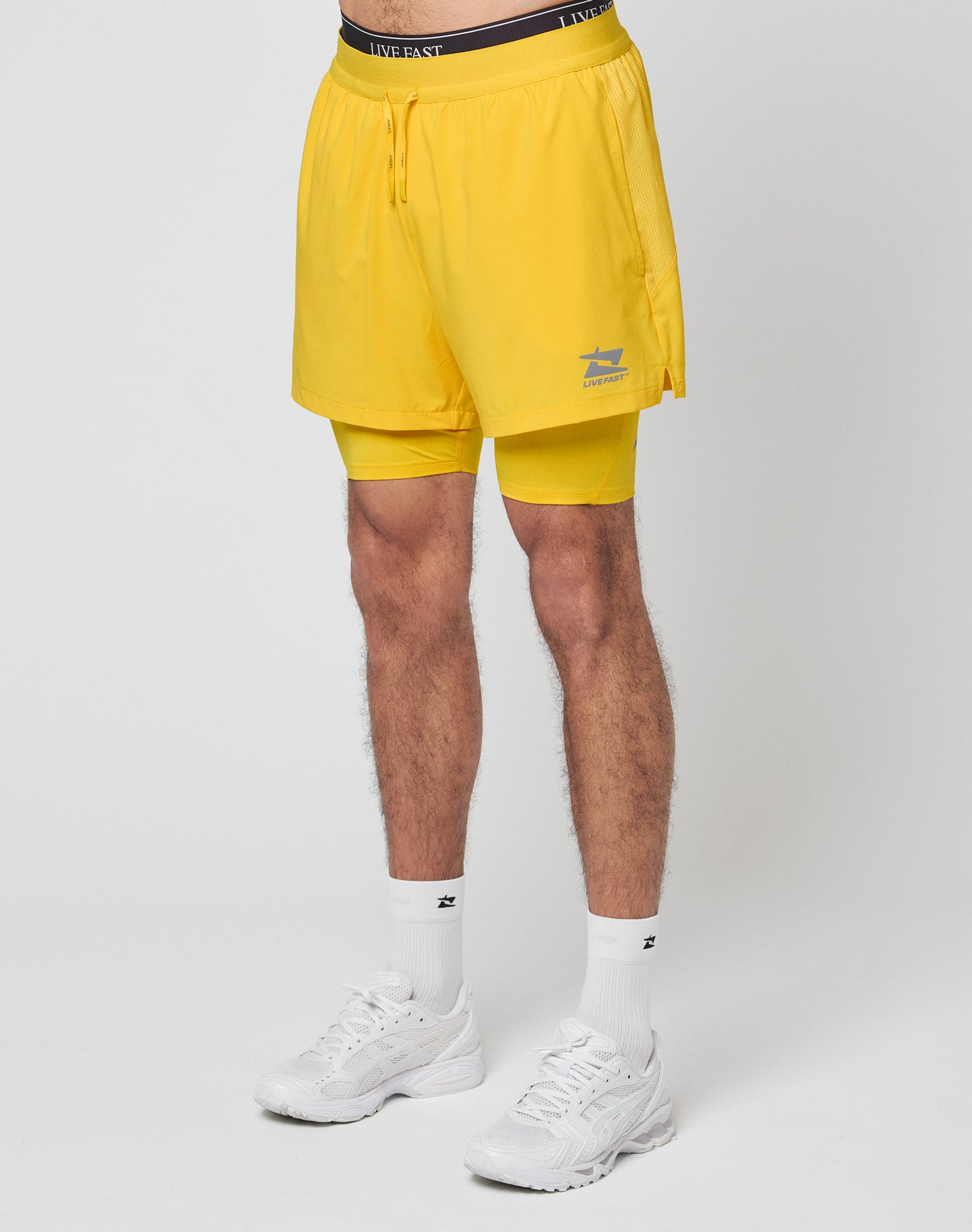A person wearing LIVE FAST DIE YOUNG Live Fast Performance Running Shorts in yellow, featuring integrated tights, along with white socks and sneakers stands against a plain white background. Only the lower body is visible.