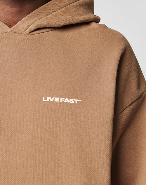 A close-up of the Live Fast Plain Hooded by LIVE FAST DIE YOUNG in tan, oversize fit, with LIVE FAST™ in white on the chest. Only part of the wearer’s shoulder, neck, and hood can be seen.