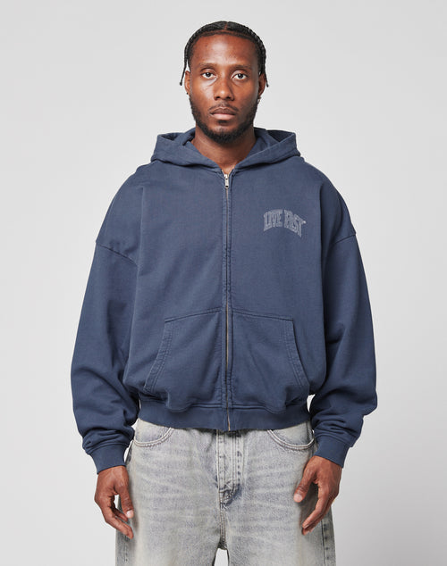 A man faces the camera in a LIVE FAST DIE YOUNG Live Fast Berlin Hooded Zip, an oversize navy hoodie with LIVE FAST text, and light gray cotton jeans, set against a plain light background.