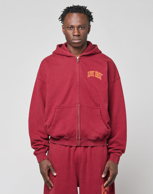 A person faces forward against a plain light gray background, wearing the LIVE FAST DIE YOUNG Live Fast London Hooded Zip in red. The hoodie features LIVE FAST in yellow on the left chest and is paired with matching pants.