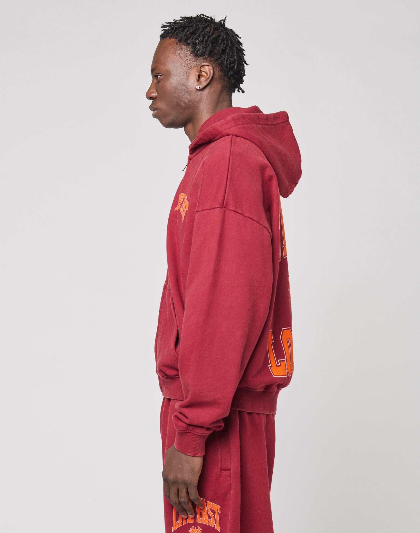 A person with short, twisted hair stands in profile against a plain background, wearing an oversized maroon LIVE FAST DIE YOUNG Live Fast London Hooded Zip and matching sweatpants with yellow lettering.