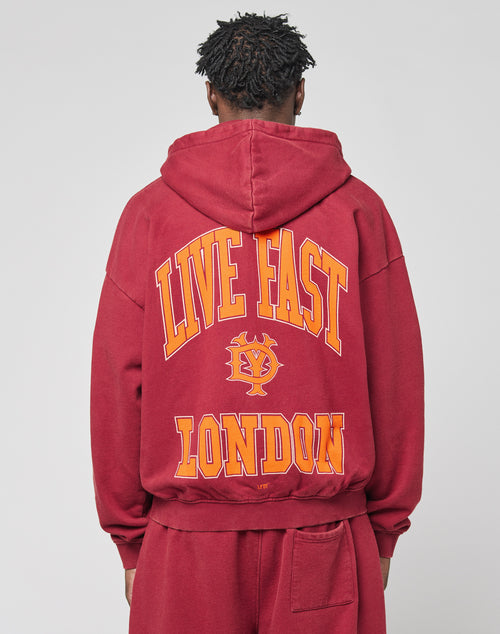 A person is seen from behind wearing an oversize fit maroon Live Fast London Hooded Zip by LIVE FAST DIE YOUNG, featuring yellow and orange LIVE FAST LONDON text and a small central graphic.