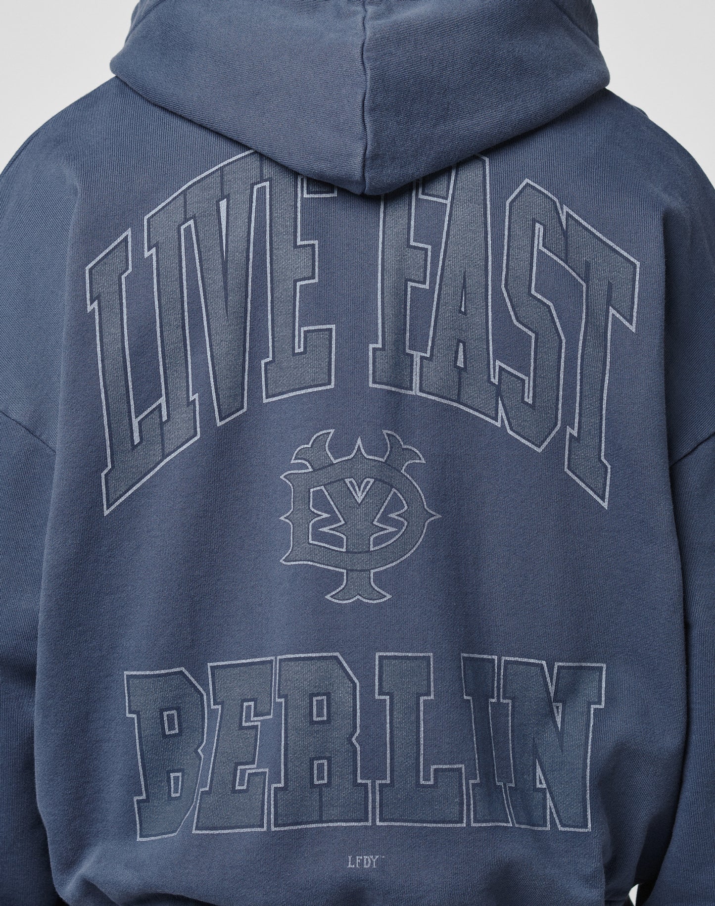 A person wearing the LIVE FAST DIE YOUNG Live Fast Berlin Hooded Zip in blue Baumwolle, oversize fit, with a vintage wash, bold LIVE FAST and BERLIN text plus graphic emblem and small LFDFY on the back.