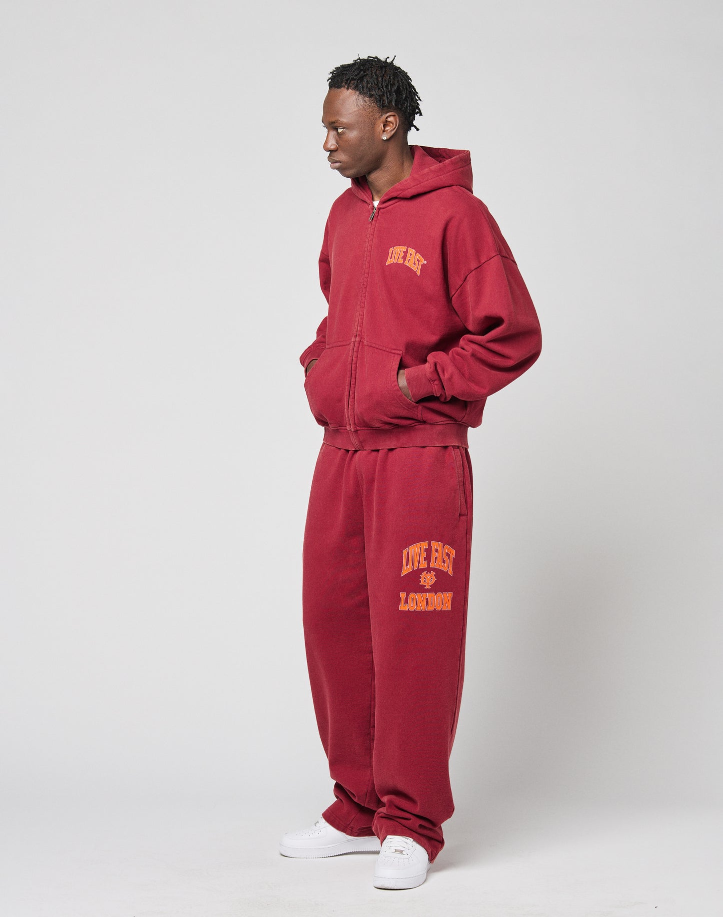 A man stands against a plain background wearing the LIVE FAST DIE YOUNG āLive Fast London Hooded Zipā and matching maroon sweatpants with yellow front and back prints, made from 100% cotton, hands in pockets, styled with white sneakers.